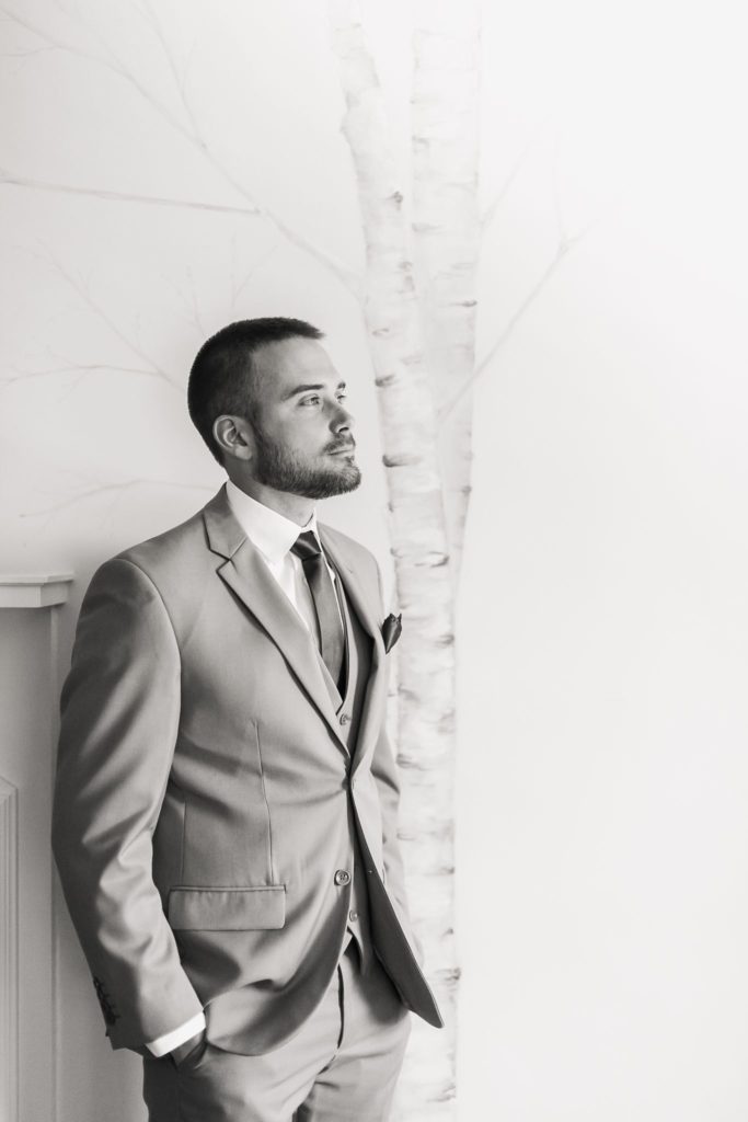 Groom in black and white standing in front of fireplace