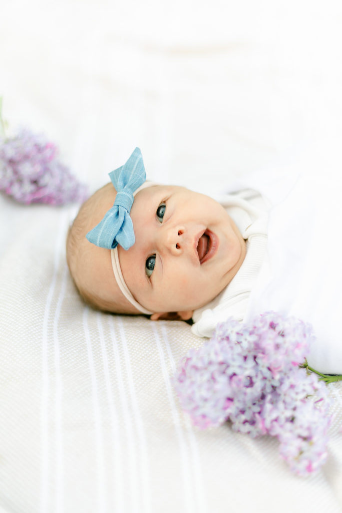 A smiling newborn baby laying on a blanket, surrounded by purple lilacs.