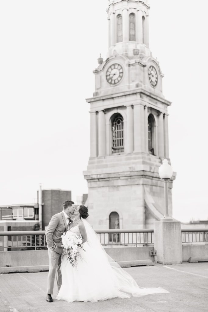 A black and white image of a wedding couple kissing on a rooftop, with skyline views in the background.