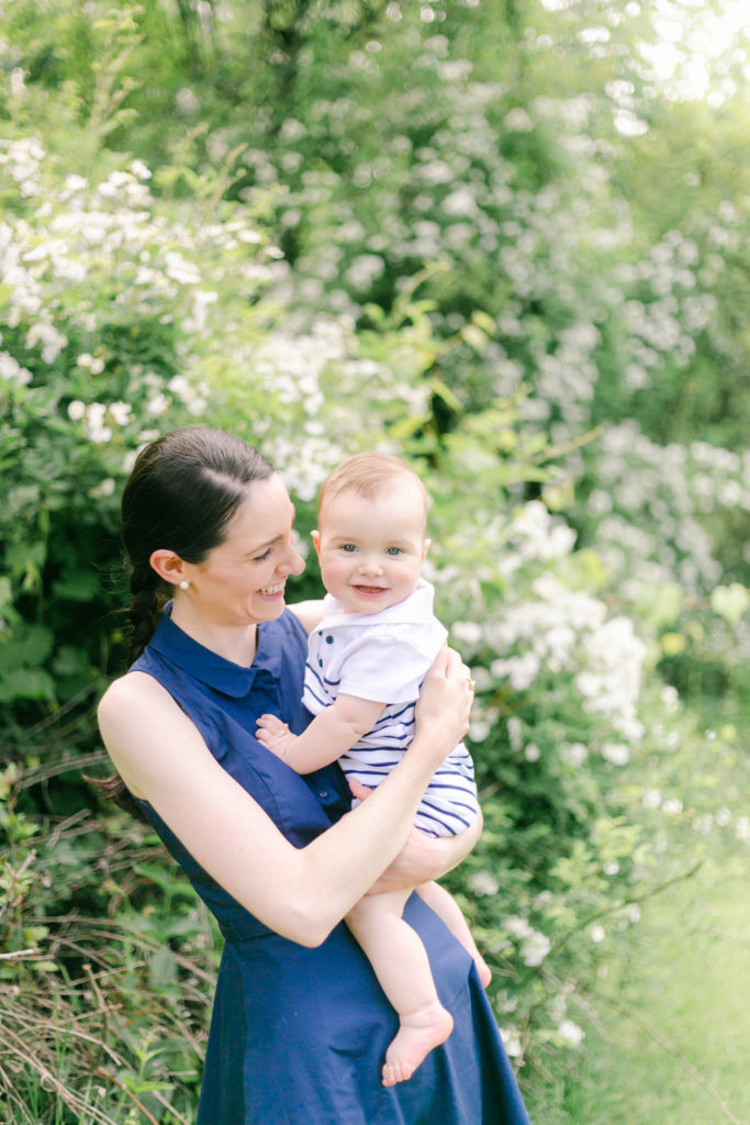 A mother in a blue dress, holding her baby in front of a blooming white rose bush.