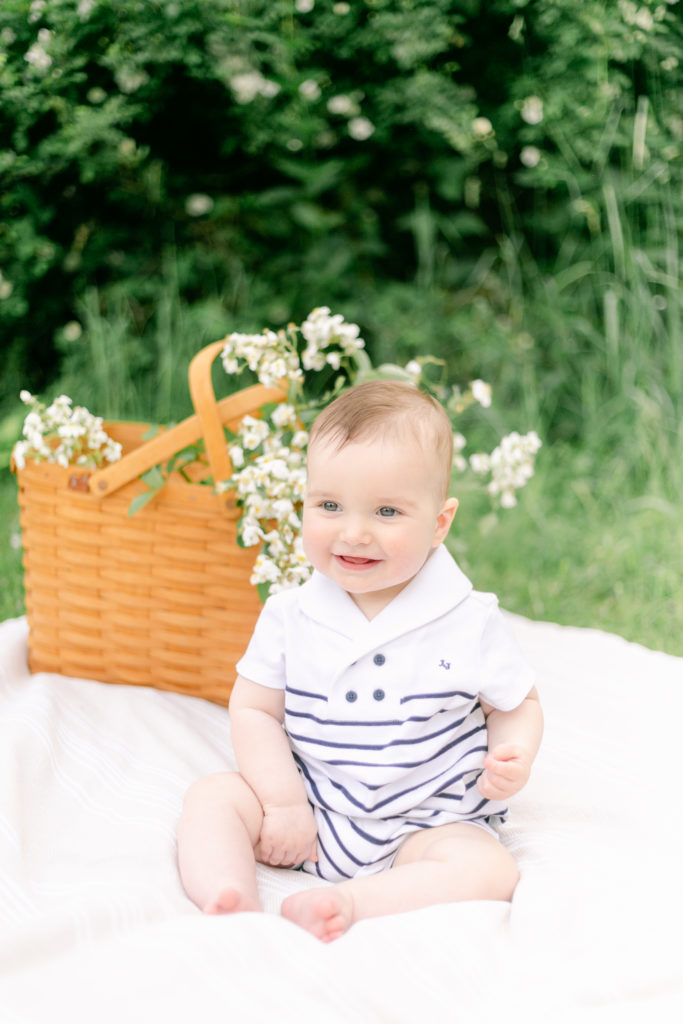 A nine month old baby sitting in front of a picnic basket filled with white roses.