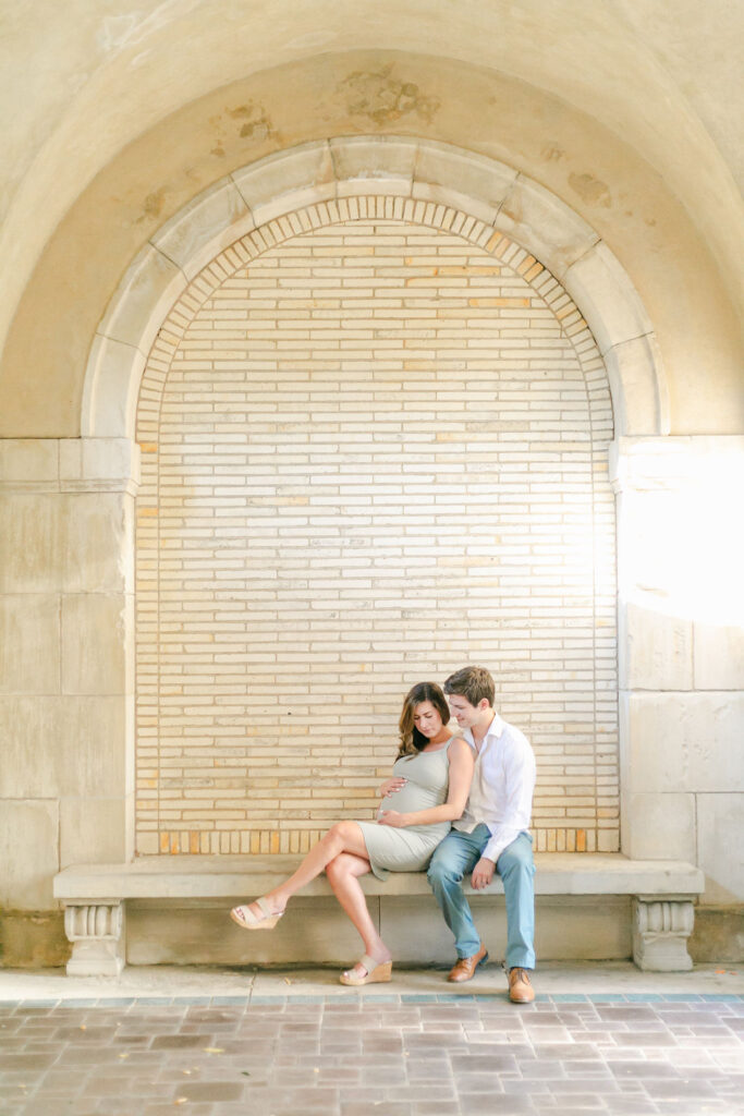 A couple sits on a bench, both looking at the woman's pregnant belly.