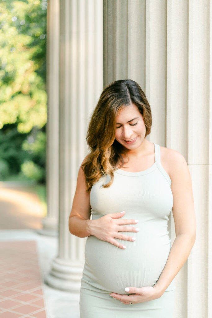 A pregnant woman hugs her belly while standing in front of an elegant row of columns.