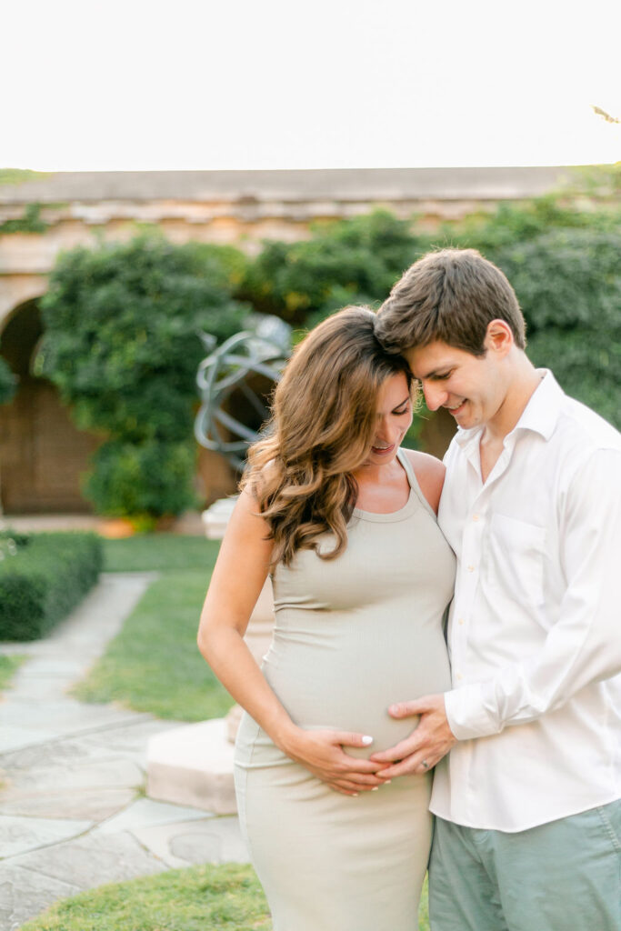 A couple caress the woman's pregnant belly while standing in an elegant garden.